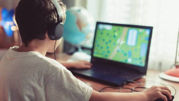 A young brunette boy in a white T-shirt wearing black headphones and playing a video game on a laptop.