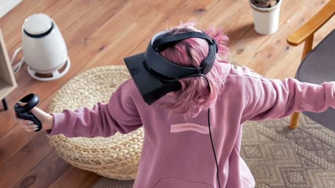 A young girl with pink hair wearing a VR headset with hand controllers and playing an active game in her living room.