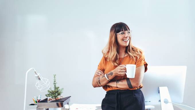 A business woman in casual wear leaning back against her desk and smiling as she crosses her arms. She has a mug in one hand.