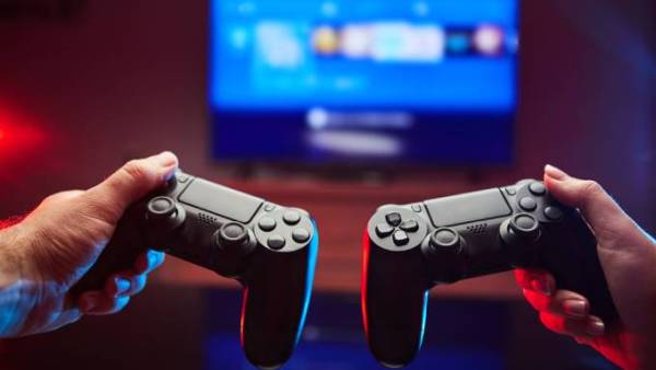 Two people holding out their video game console controllers while looking on at a blurry TV showing a console’s home page.