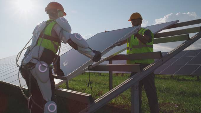 Two workers in high-visibility gear working to set up a solar panel. One is also wearing an assistive exoskeleton.