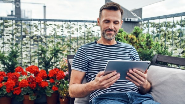 A man sitting on a patio lounge while smiling down at a digital tablet. An array of flowers and plants are behind him.