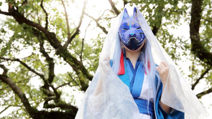A cosplayer at an outdoor event. They are wearing a kimono and a fox mask with a veil draped over their cosplay.