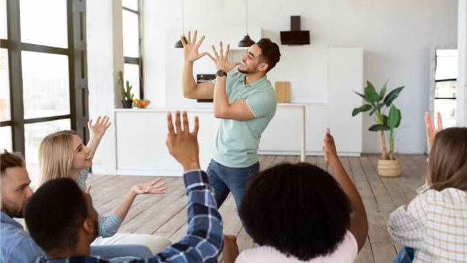 A group of young friends play charades in a modern home living room. One man mimes an action while the rest shout guesses.