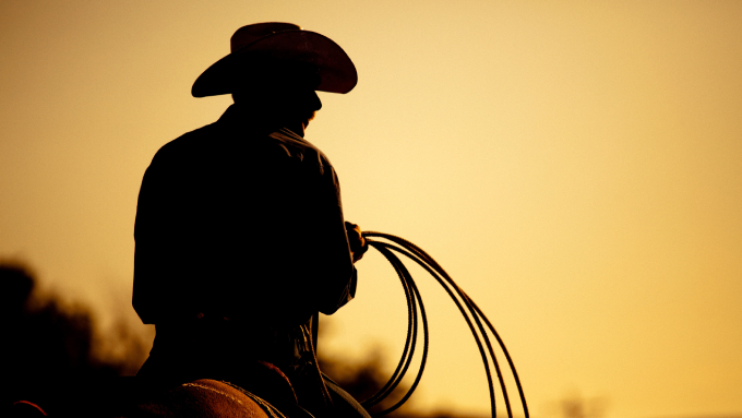 The silhouette of a cowboy wearing a cowboy hat and holding a lasso while riding a brown horse at sunset.