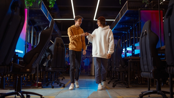 Two boys are talking and fist-bumping while walking in an aisle of a video game store with rows of computers and chairs.