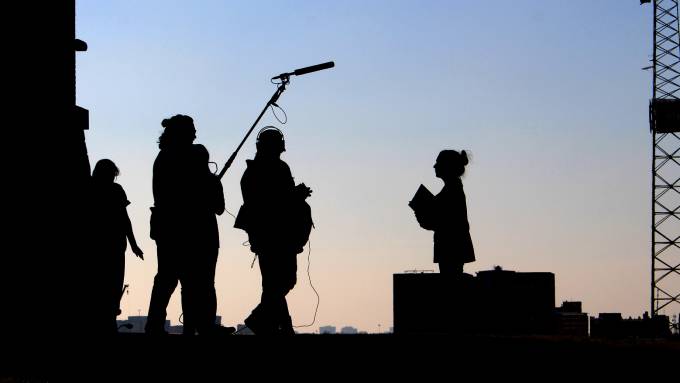 The silhouette of a film crew standing outside. Someone holds a microphone, and another has film equipment.