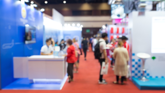 A blurred visual of a convention event with people background. The carpet is all red with colorful booths.