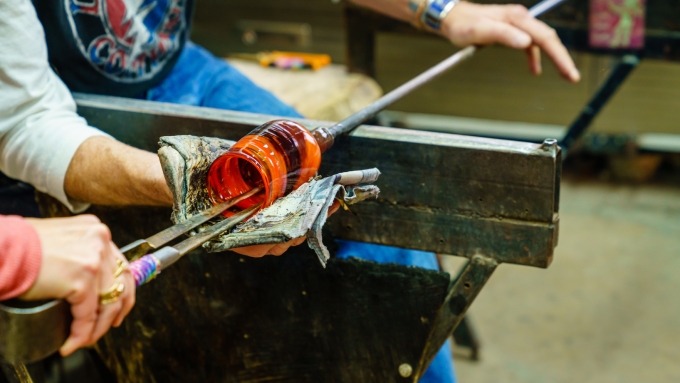 A close-up view shows two people making art with glass. One person uses tweezers on the glass while the other holds it with a rag.