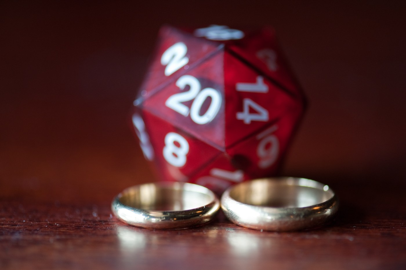 A pair of differently sized golden wedding bands in the foreground with a red D20 die in the background.