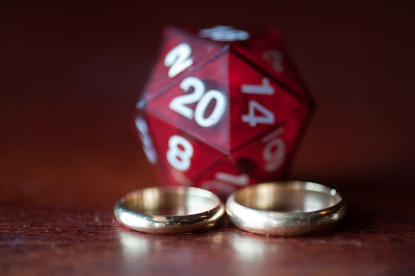 A pair of differently sized golden wedding bands in the foreground with a red D20 die in the background.