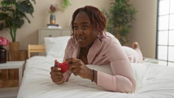 A woman in pink satin pajamas laying on your stomach in bed and playing a game with a red gaming controller.