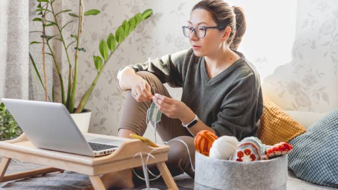 A woman sitting on the floor and knitting, her supplies next to her, as she watches something on her laptop.