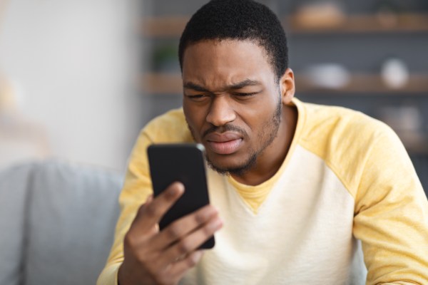 A Black man in a baseball tee, holding his phone close to his face, mistrustfully squinting at the screen.