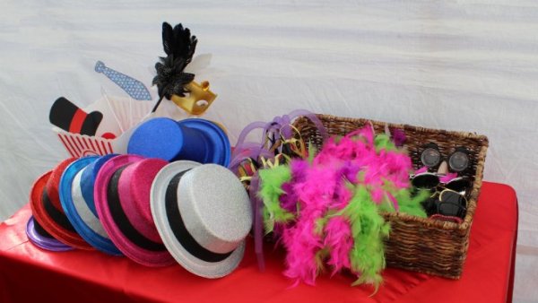 A table with a red cover contains various photo booth props, including colorful hats, sunglasses, and fuzzy boas.
