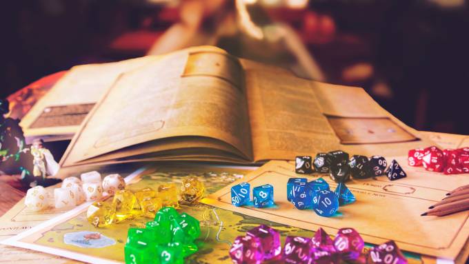 An assortment of colorful acrylic game dice alongside painted miniatures in front of several open tabletop game books.