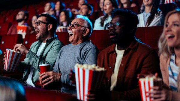 Many people sitting in red movie theater seats and smiling widely as they look at the screen. They are holding snack and drinks.