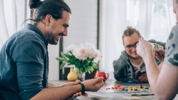 A smiling man adjusting his watch as he looks down at the cards from a colorful boardgame. His friends sit with him at the table.