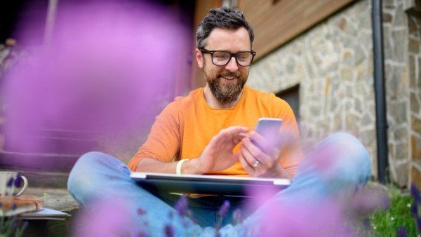 A smiling man in an orange shirt sitting on the ground outside with a laptop in his lap and a phone in hand.