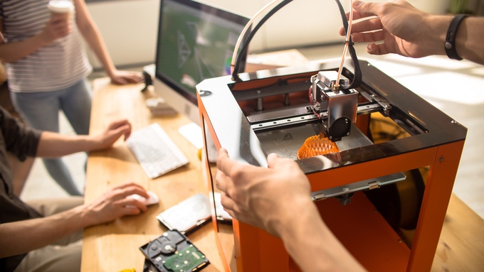 Three people in a design studio review a design on their computer while it prints from their 3D printer.