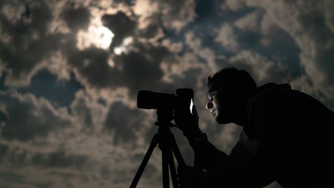 A photographer using his camera in low lighting outside. The clouds cover the moon as he looks at the camera's screen.