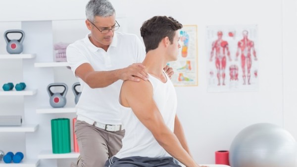 A young man wearing a tank top sitting on a table in a clinic as a chiropractic makes adjustments to his back.