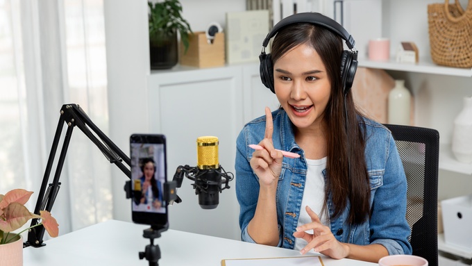 A woman talking to a mounted mic and a phone that's filming her. A piece of paper and a pink mug are on top of the table.