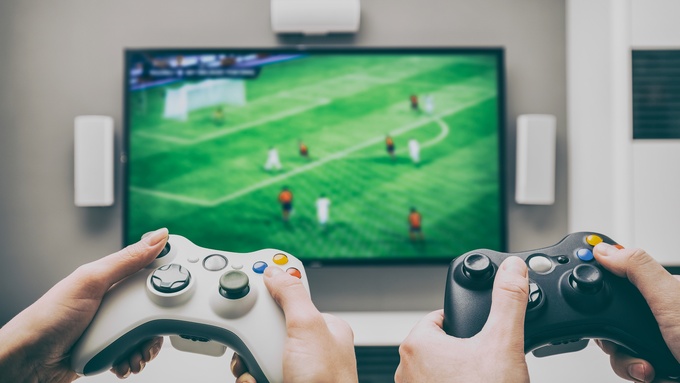 Two people play a soccer video game on a flat-screen TV. One holds a white controller, and the other holds a black one.