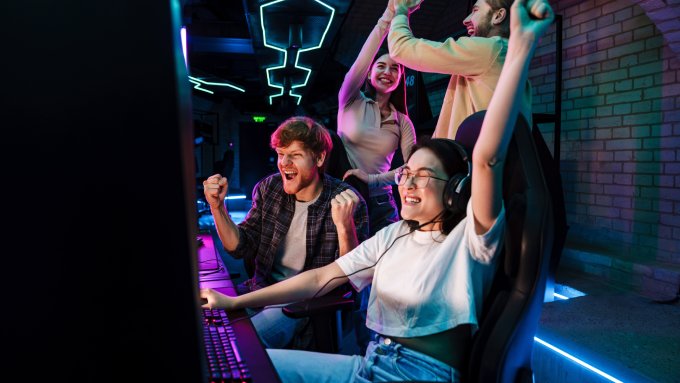 A group of people sit around a gaming laptop. They all cheer and high-five each other.