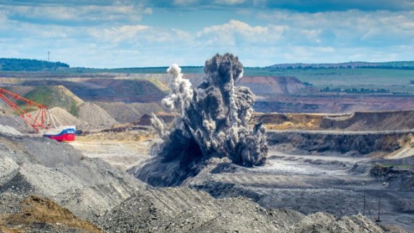 A large, cloudy, gray explosion occurring in an open pit within an expansive rock mining quarry.