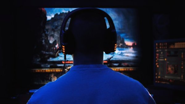 The back of a man's head as he plays video games on his PC. The light of the screen illuminates the area around him.