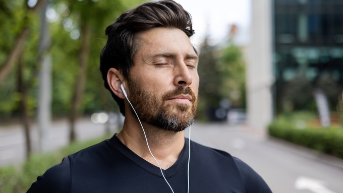 A person with a beard standing in public and closing his eyes as he listens to audio through white ear buds.