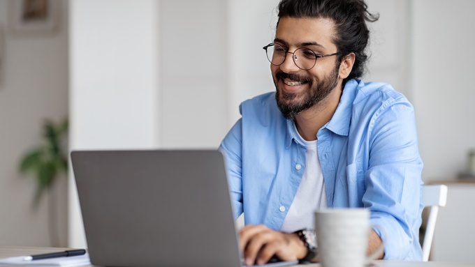 A man wearing a blue shirt and black glasses sits at a white desk, typing on a silver laptop. A coffee cup is next to him.