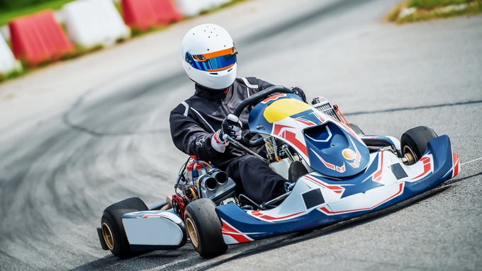A person is driving a blue, white, and red go-kart wearing a white helmet and black suit on an outside go-kart track.