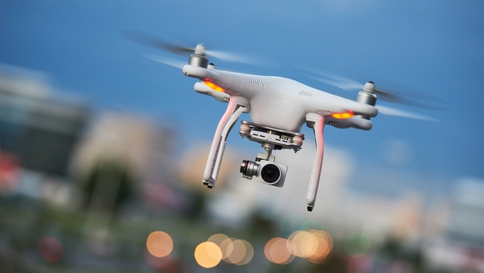 A close-up of a drone with a camera in mid-flight. The background is blurred with orbs of light present.