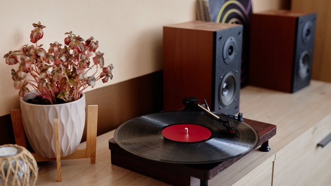 A turntable with a spinning record rests on a table. There are two speakers to the right and a plant to the left of the turntable.