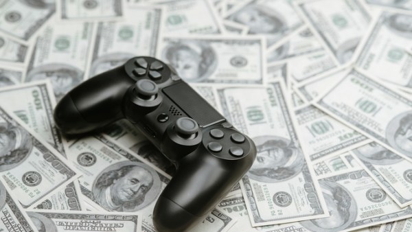 A black controller used for playing video games sits on top of a pile of several one-hundred-dollar bills.