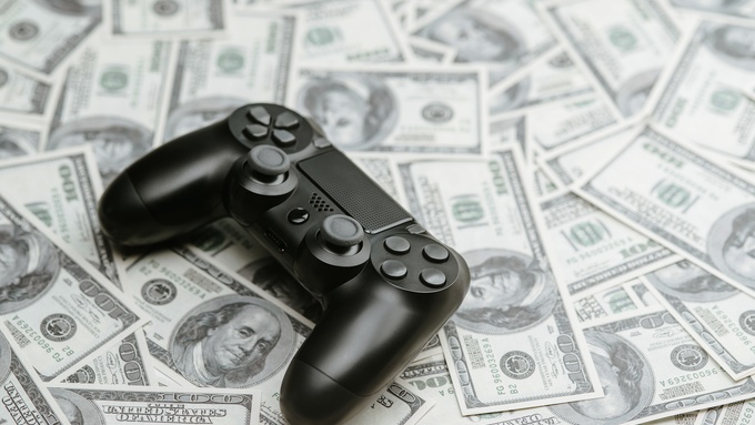 A black controller used for playing video games sits on top of a pile of several one-hundred-dollar bills.