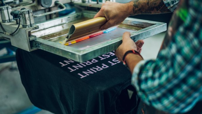 A worker with tattoos applying a rainbow of ink to a printing screen. The words on the black T-shirt are pink.