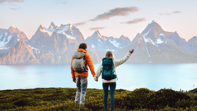 Two people wearing hiking backpacks standing on a cliff overlooking water. There are beautiful mountains in the distance.