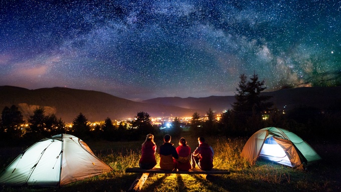Friends sitting together while camping under a star-filled night sky, with two tents set up beside them in the background.