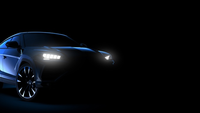 The front view of a dark blue sports car with its high-beam light on and parked against a dark background.