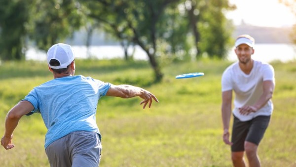Two men wearing white hats, T-shirts, and shorts, smile and play frisbee in a park near trees and a lake.