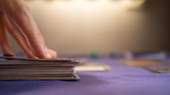 A hand lifting a card from the top of a playing card deck. Other cards and dice are visible in the background.