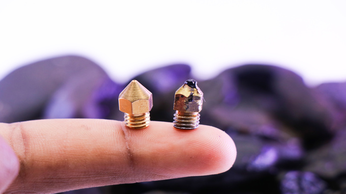A close-up view shows two gold 3D printer nozzles sitting on a finger. One is new and one is damaged.