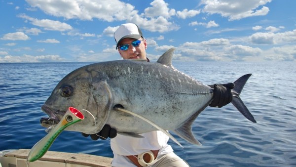 A man wearing a white hat standing on a boat in the ocean while holding a large fish that he just caught.