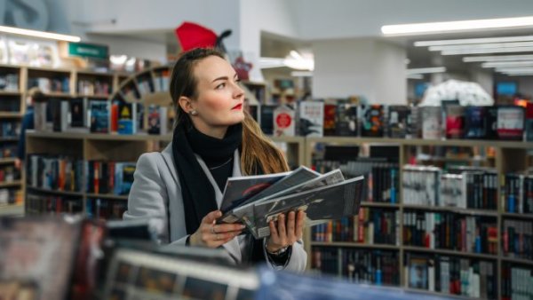 A woman in a pop culture store chooses from four comic books as she walks between two shelves filled with options.