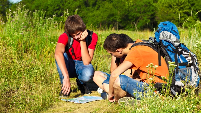 Three people wearing camping backpacks sit around a map on a sunny day, planning their route along a trail in the outdoors.