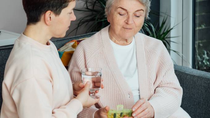 An older parent examines their pill container while their adult child sits close by with a small glass of water.