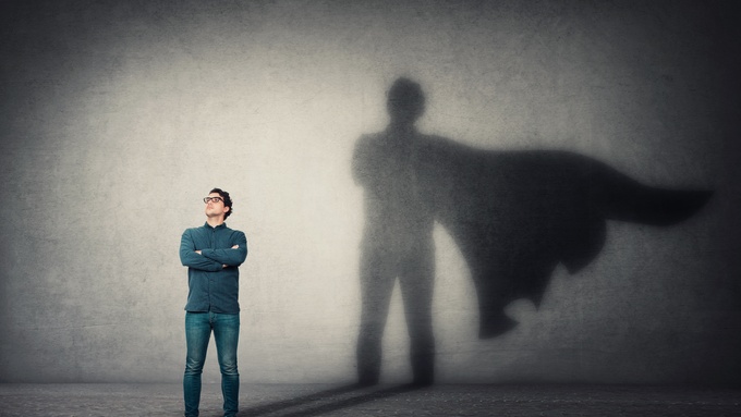 A man standing with his arms crossed in front of a gray wall. His shadow is very large, with a cape flowing behind him.